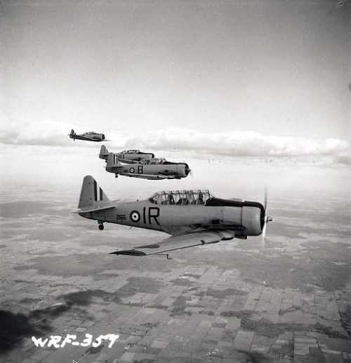 Black and white photograph. Four aircraft fly through the air in a line beside each other. On the ground, the patchwork appearance of fields.